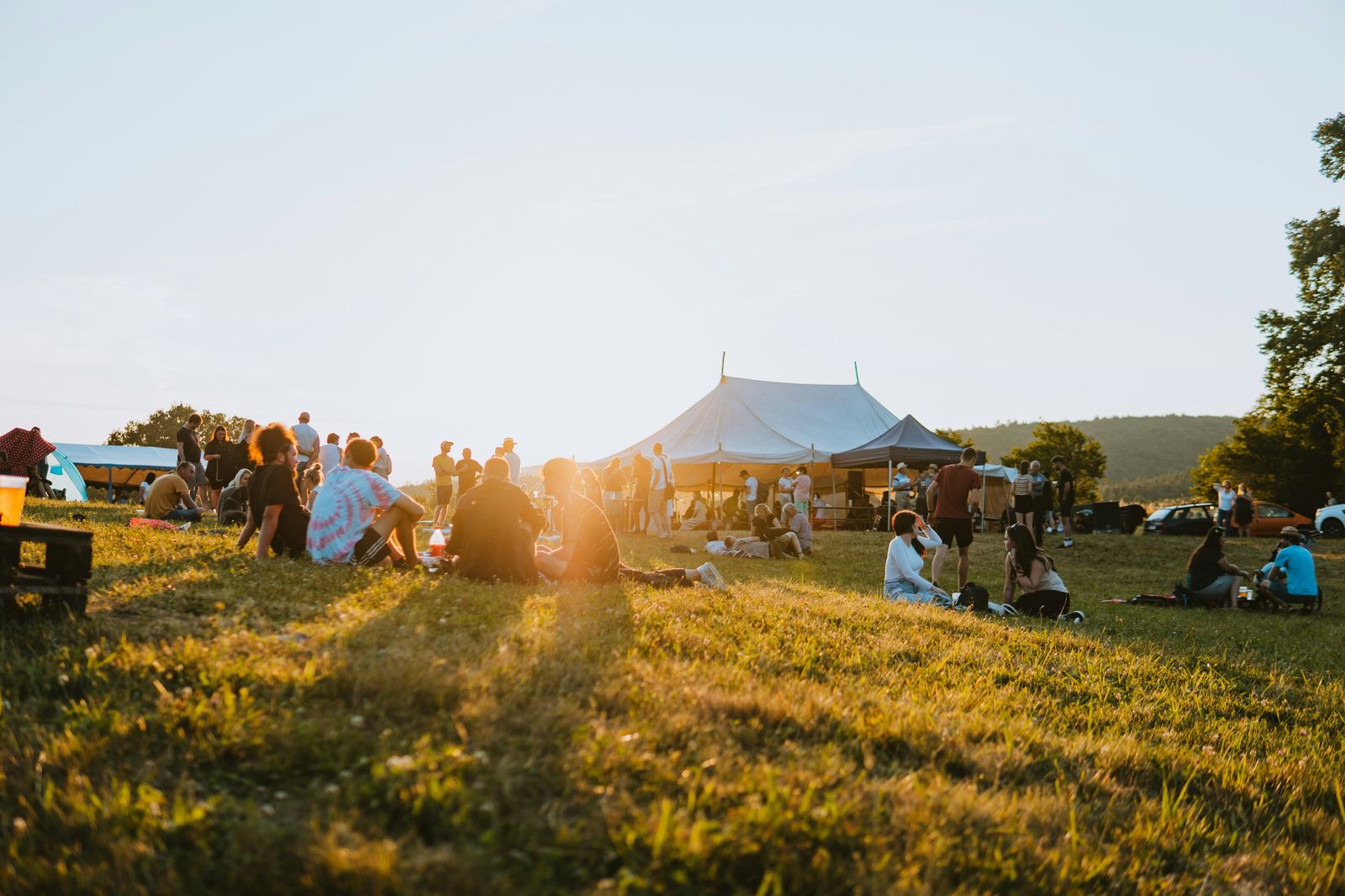 Small festival in a field at sunset, with groups sitting on the grass in front of a large marquee tent