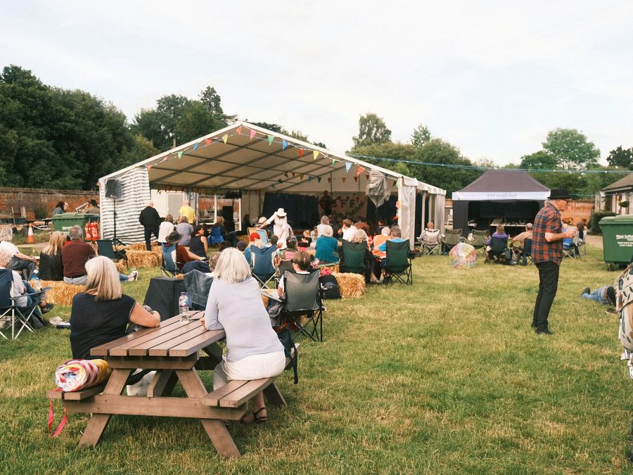 Community festival audience in camping chairs and on hay bales, watching a band under a marquee