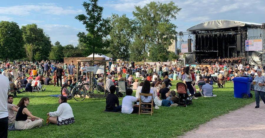 Crowd sitting on the grass in a park in front of a large outdoor concert stage