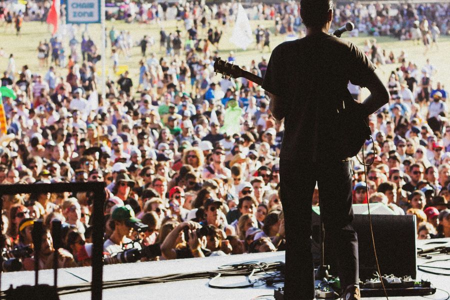 View from behind a performer on stage, looking out over a large festival crowd in a park