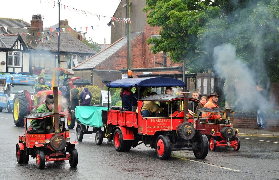 Miniature steam engines carrying passengers along a closed village street decorated with bunting